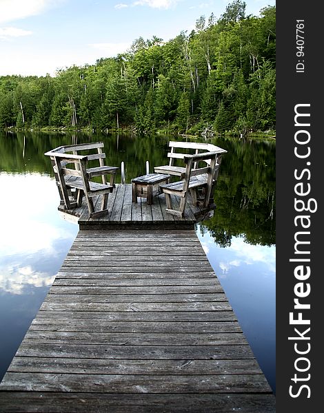 View of a wooden dock at the lake, with trees in the background and a reflection of the trees in the water.