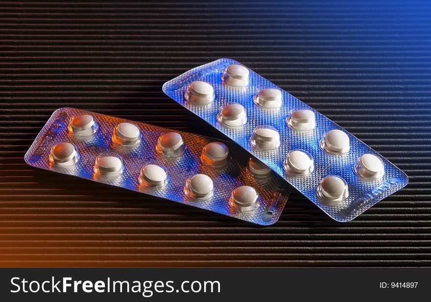 Two pill packages lit by orange and blue lights on black background. Two pill packages lit by orange and blue lights on black background