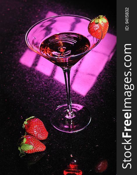 A martini glass with red liquid splashing up in center and a strawberry placed on side with two strawberries next to glass. A martini glass with red liquid splashing up in center and a strawberry placed on side with two strawberries next to glass