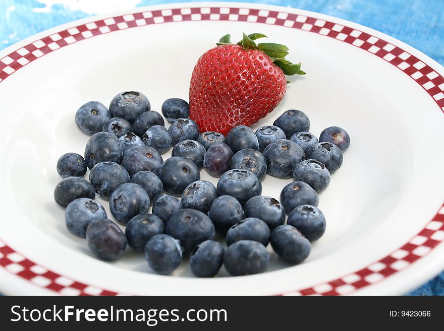 Many blueberries and a single strawberry on a plate in the sun. Many blueberries and a single strawberry on a plate in the sun