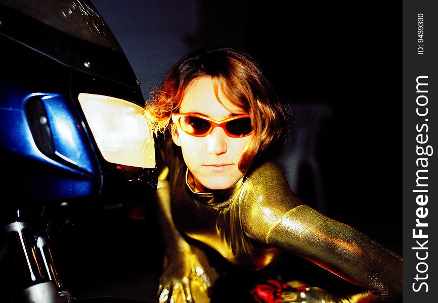 A young woman wearing sunglasses dressed in a yellow jump suite with her face near the motorcycle light.