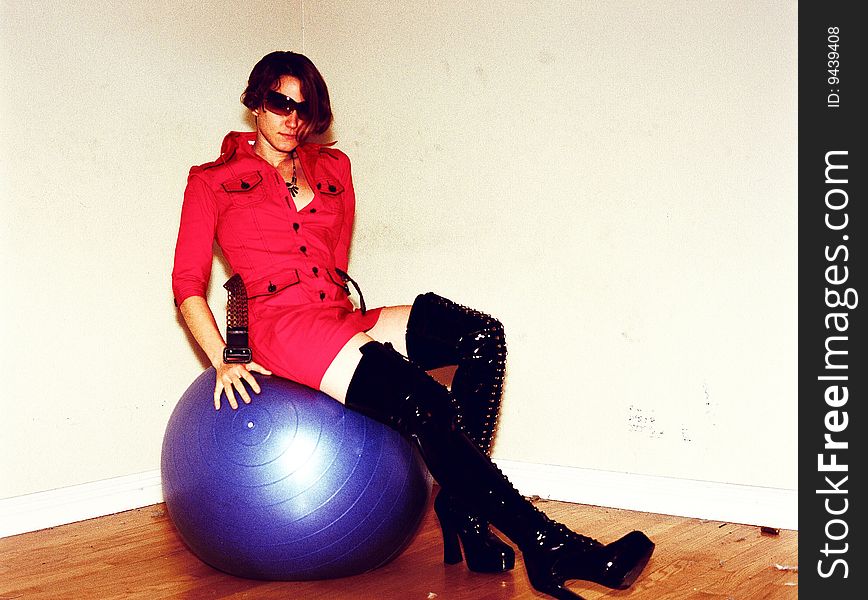 A young blonde woman wearing a red jump suite and long black boots who is sitting on top of a big blue exercise ball. A young blonde woman wearing a red jump suite and long black boots who is sitting on top of a big blue exercise ball.