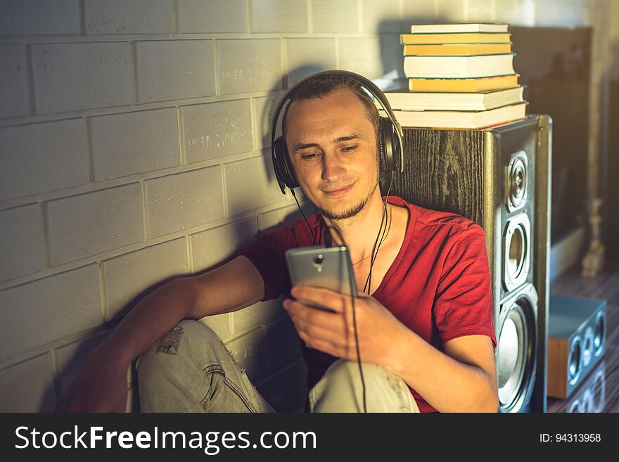 Attractive young man listening to the audio book in the headphones with a mobile phone in a modern interior with books on the background of multimedia and large speakers. The concept of technology education a positive lifestyle. Attractive young man listening to the audio book in the headphones with a mobile phone in a modern interior with books on the background of multimedia and large speakers. The concept of technology education a positive lifestyle