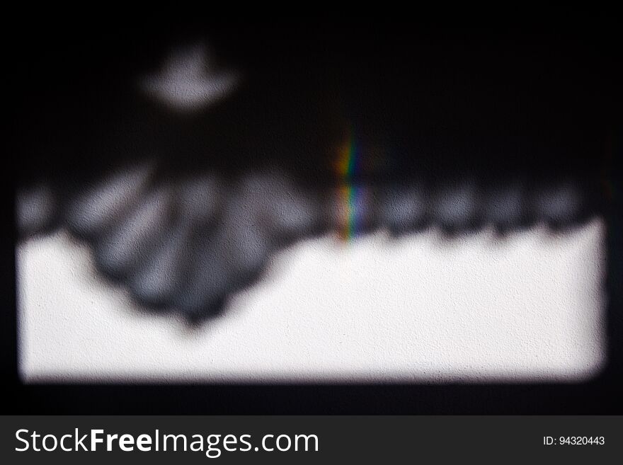 A shadow of a window curtain on a white wall, with a small rainbow generated by the sun rays passing through the window. A shadow of a window curtain on a white wall, with a small rainbow generated by the sun rays passing through the window.