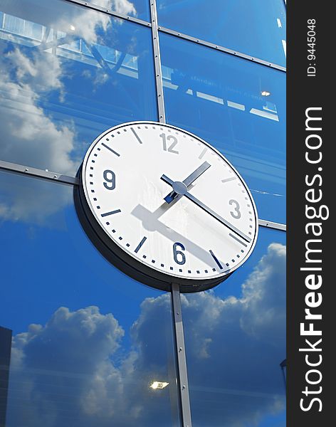 Picture of a clock on glass wall reflecting clouds and blue sky