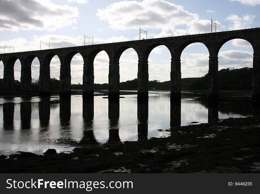 A silhouette of the Royal Broder Railway Bridge at Berwick Upon Tweed in Britain, shot against a cloudy blue sky and the piers reflecting in the water. A silhouette of the Royal Broder Railway Bridge at Berwick Upon Tweed in Britain, shot against a cloudy blue sky and the piers reflecting in the water.