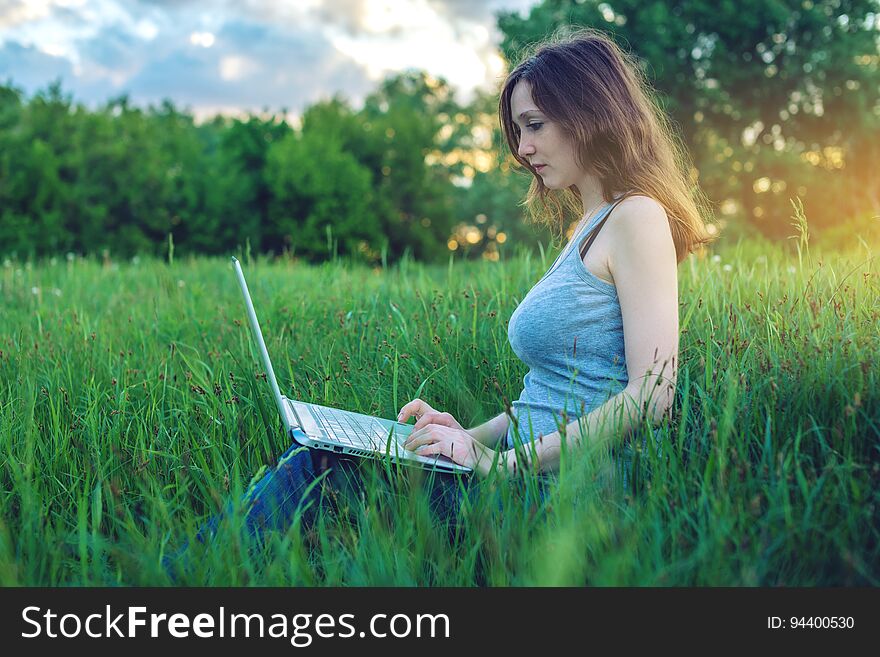 Woman sitting on a green meadow with trees on the background of sunset with clouds. Working or studying on laptop