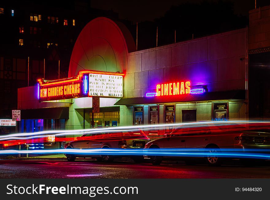 A movie theatre illuminated at night with it's neon signs and light trails from passing cars.