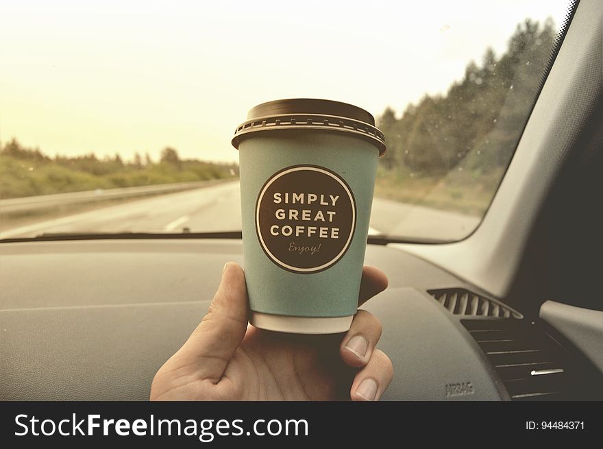 Hand holding disposable coffee cup inside car on road. Hand holding disposable coffee cup inside car on road.