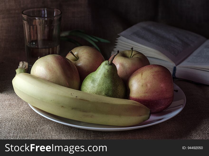 Still life with apples pear and banana in the background is a glass with a book