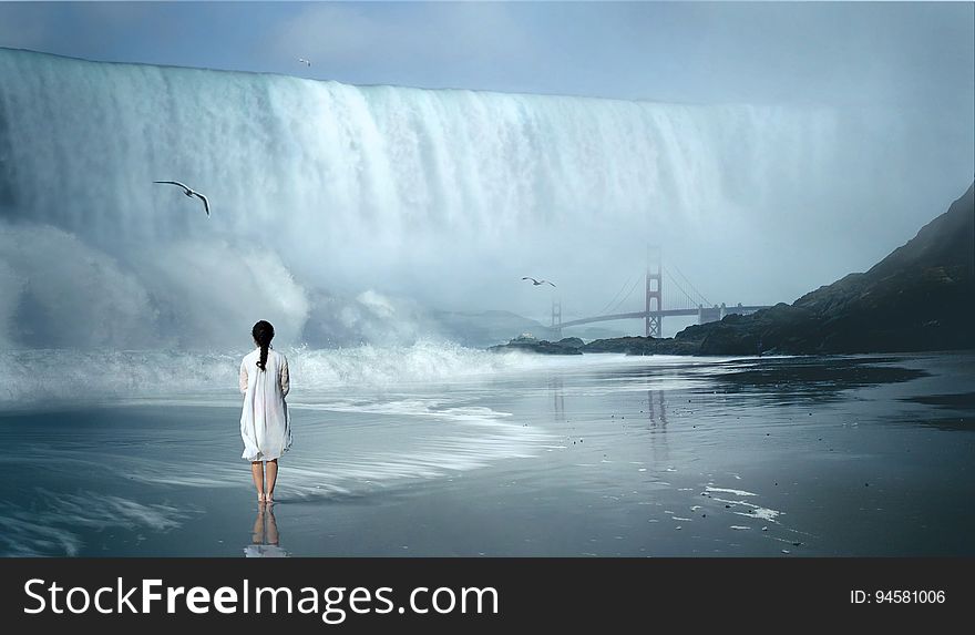 Young woman in white dress standing in water looking towards a dramatic waterfall with seagull overhead and suspension bridge in the distance (suggesting a composite image). Young woman in white dress standing in water looking towards a dramatic waterfall with seagull overhead and suspension bridge in the distance (suggesting a composite image).