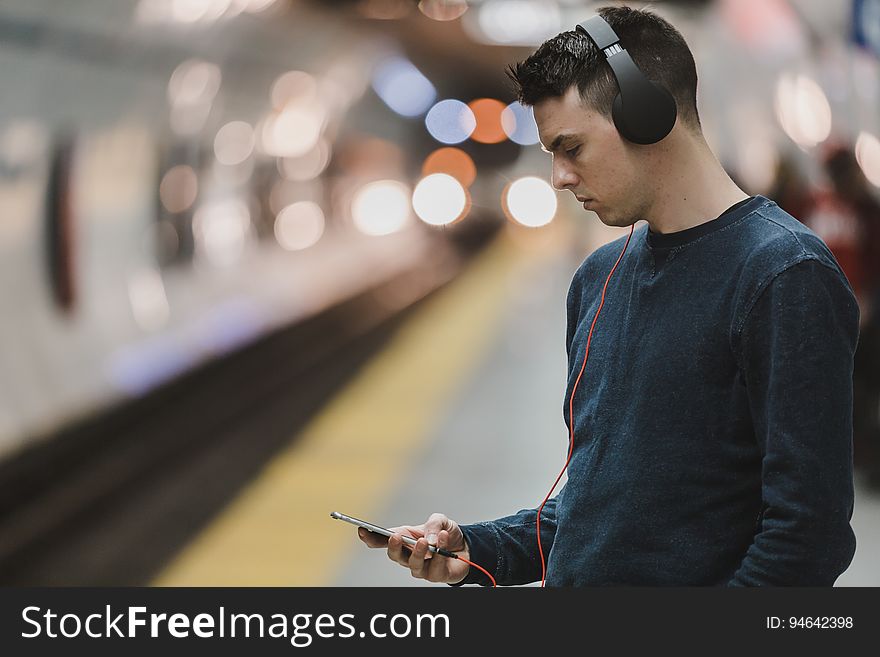 A man waiting for a train or a subway looking at his phone with headphones on. A man waiting for a train or a subway looking at his phone with headphones on.