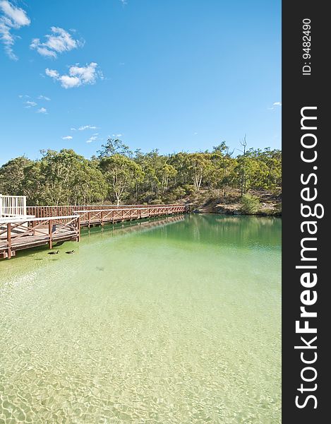 A wooden bridge beside a beautiful clear crystal lake, surrounded by vegetations and against the blue sky. A wooden bridge beside a beautiful clear crystal lake, surrounded by vegetations and against the blue sky.