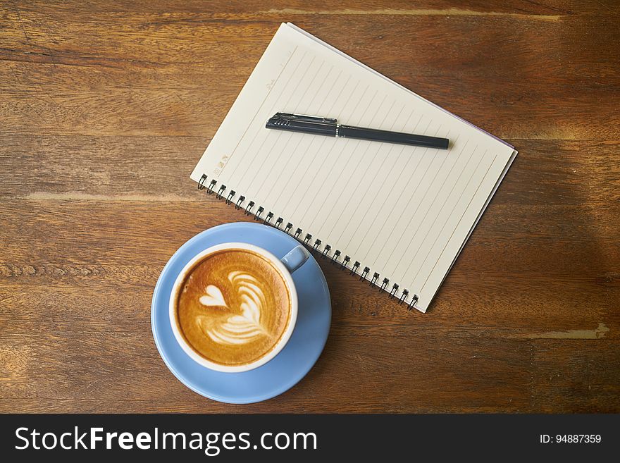 Overhead view of coffee on wooden table with decorative heart shaped next to notepad and pen. Overhead view of coffee on wooden table with decorative heart shaped next to notepad and pen.