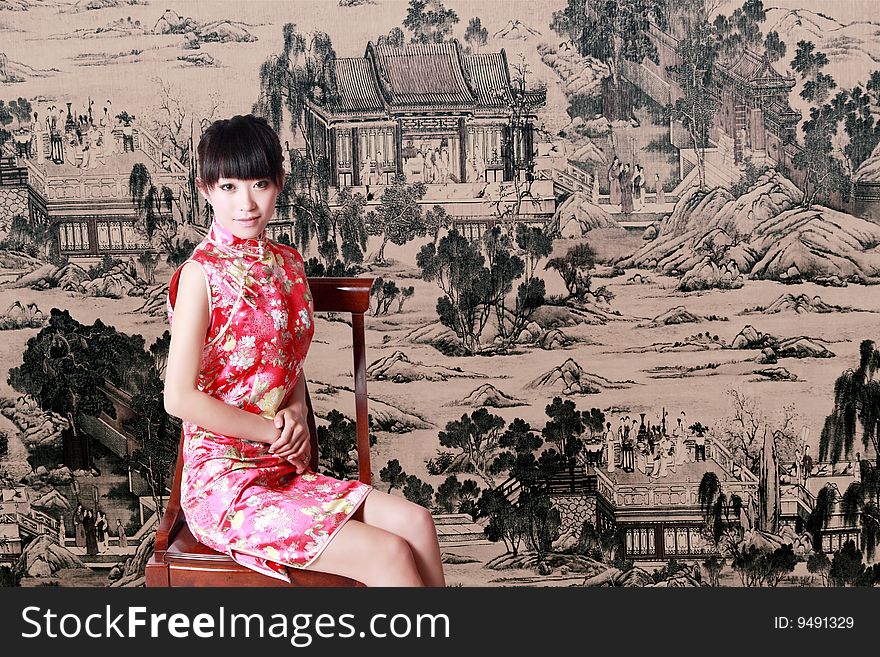 A Chinese girl in traditional dress sits in front of a Chinese old painting.