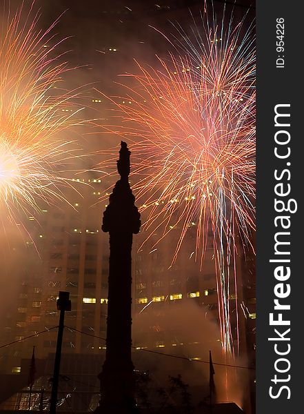 A fireworks display in a downtown setting with a skyscraper as the background and a memorial in the foreground. A fireworks display in a downtown setting with a skyscraper as the background and a memorial in the foreground