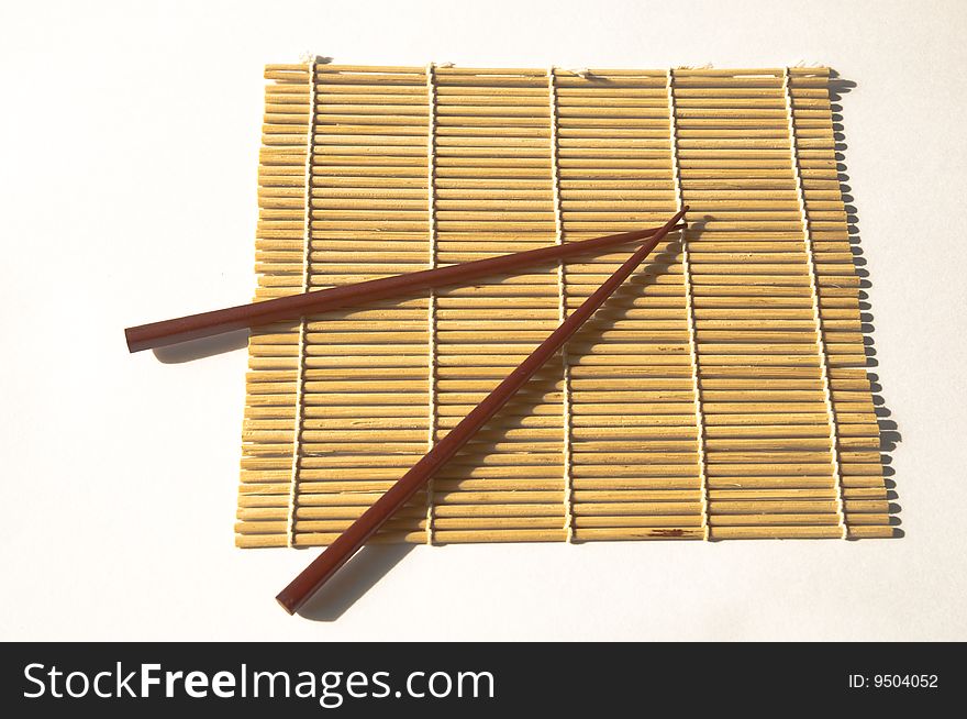 A Pair or square wooden chopsticks on a bamboo maki rolling mat on a white background. A Pair or square wooden chopsticks on a bamboo maki rolling mat on a white background