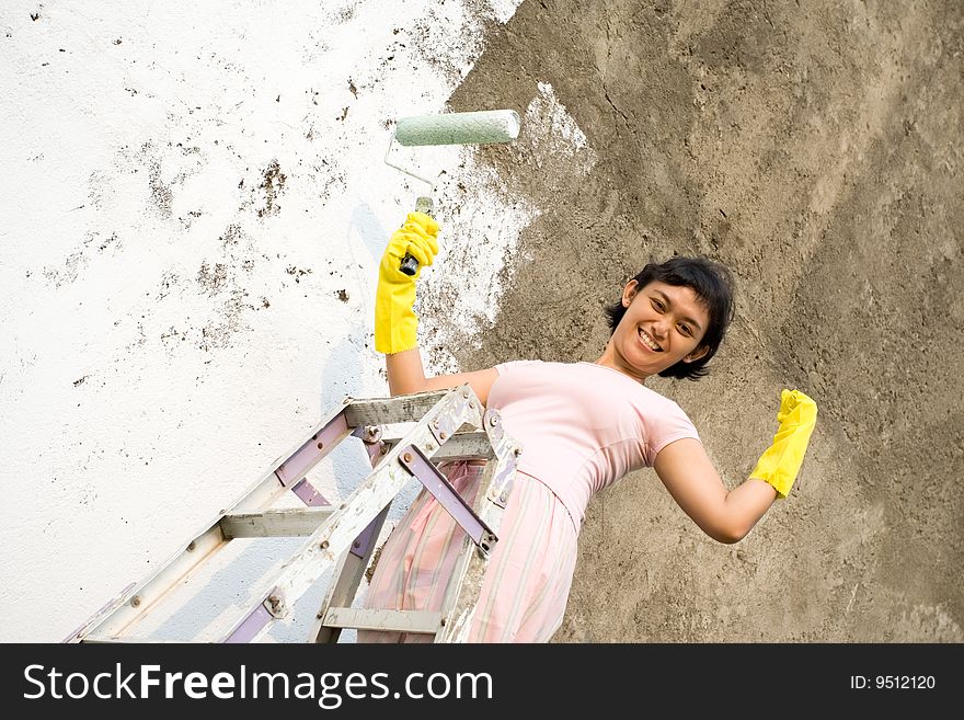 Cheerful asian woman showing her strength, capacity and capability. painting exterior wall of her house by herself with a white colored paint, using a ladder, roller and gloves on hands. Cheerful asian woman showing her strength, capacity and capability. painting exterior wall of her house by herself with a white colored paint, using a ladder, roller and gloves on hands