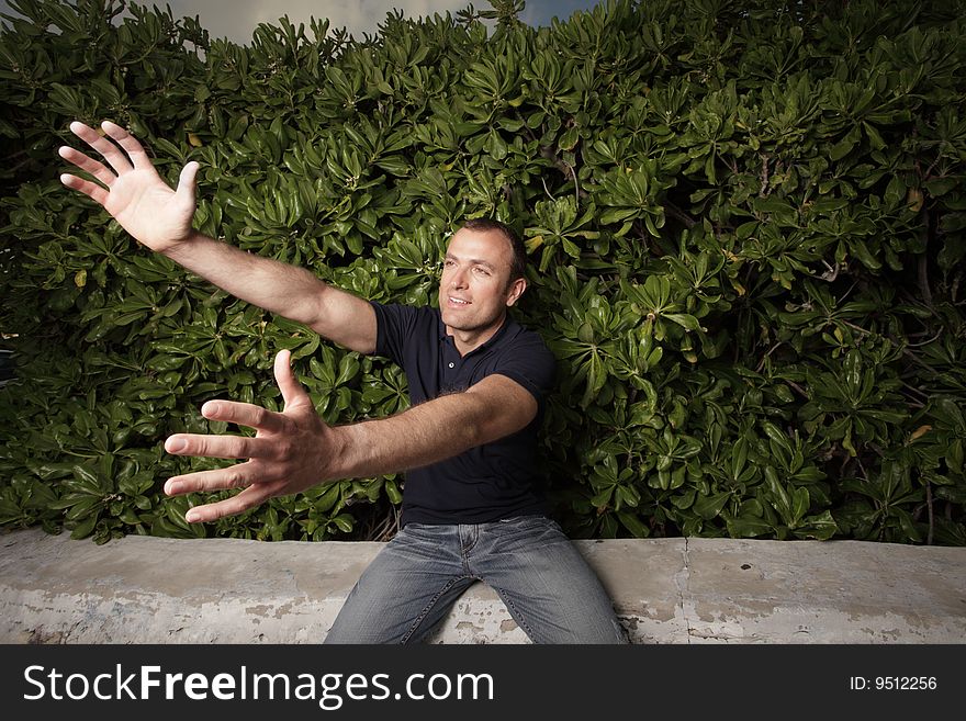 Young man sitting on a ledge by bushes and reaching out his arms