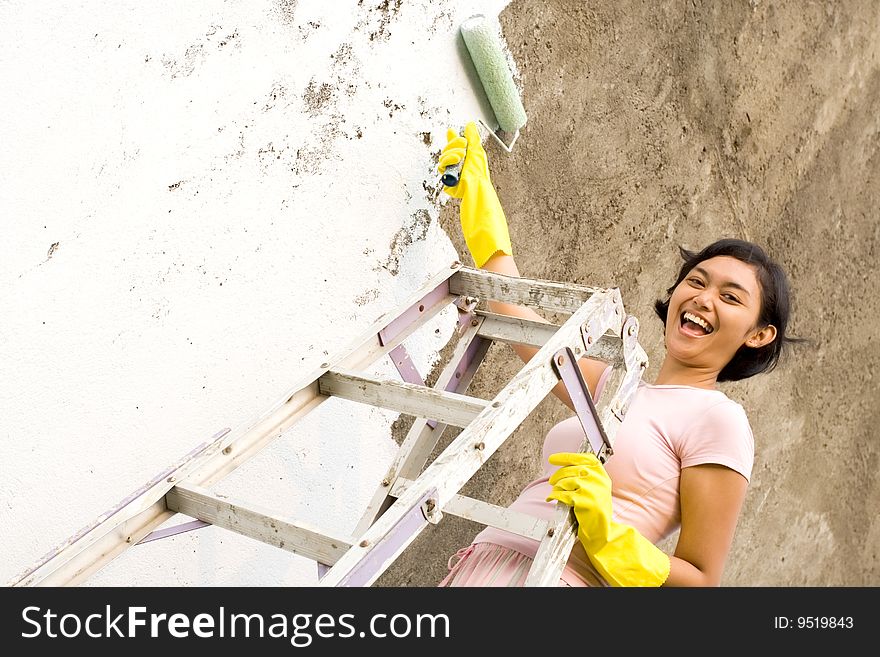 Cheerful asian young woman standing on a ladder, painting exterior wall of her house with a white colored paint, using a roller and gloves on hands. Cheerful asian young woman standing on a ladder, painting exterior wall of her house with a white colored paint, using a roller and gloves on hands