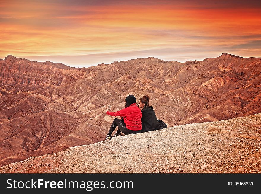 2 Woman Seating On The Mountain Cliff During Golden Hour