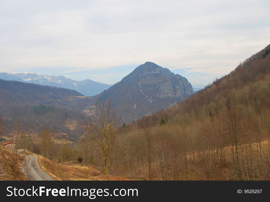 Italy. The Alpes. Early spring. Still there is no green grass. On trees there is no foliage. At mountain tops the snow has not thawn. The nature waits for present heat. Cloudy day. A kind on a valley, mountains from a slope of a hill. At the left – the road going downwards, country economic construction. Italy. The Alpes. Early spring. Still there is no green grass. On trees there is no foliage. At mountain tops the snow has not thawn. The nature waits for present heat. Cloudy day. A kind on a valley, mountains from a slope of a hill. At the left – the road going downwards, country economic construction.