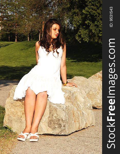 An young lovely girl with long brunette hair sitting on a big rock in the park
in sunset with an white dress. An young lovely girl with long brunette hair sitting on a big rock in the park
in sunset with an white dress.
