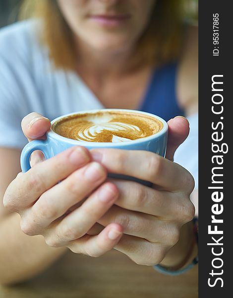 Closeup of two hands of a woman wrapped around a blue cup or mug of coffee (latte) with a partial blurred face behind. Closeup of two hands of a woman wrapped around a blue cup or mug of coffee (latte) with a partial blurred face behind.