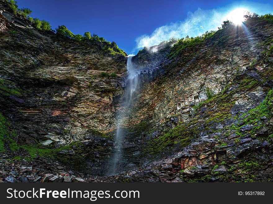 An impressive waterfall down a rocky cliff with a fringe of trees on top and green moss down below, blue sky and white cloud above.