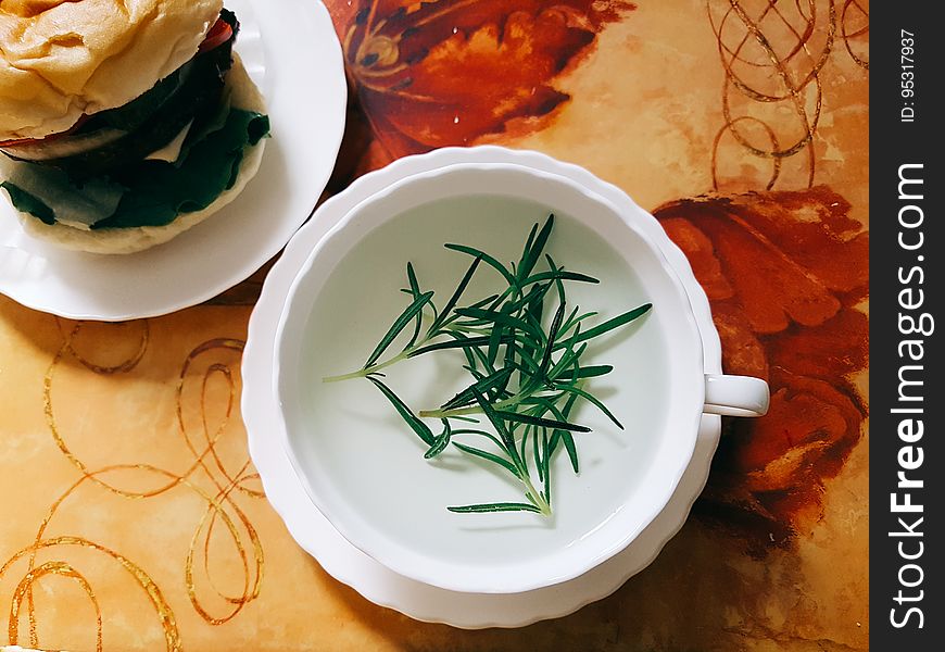 Cup and saucer with green herbs in clear broth with hamburger on white china plate. Cup and saucer with green herbs in clear broth with hamburger on white china plate.