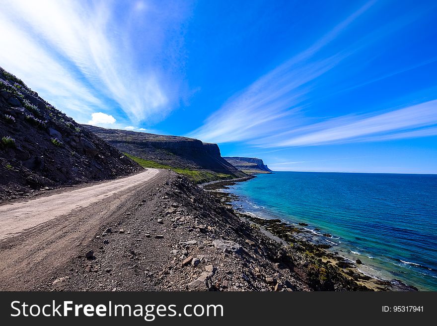 Rough coast road beside an aquamarine sea with dramatic sky with clouds in divergent white streaks. Rough coast road beside an aquamarine sea with dramatic sky with clouds in divergent white streaks.
