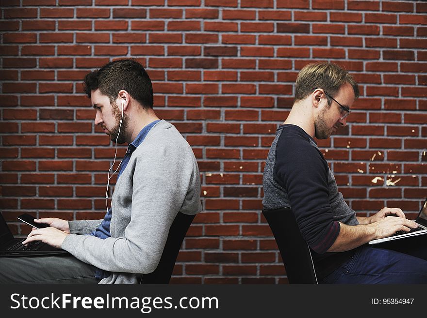 A pair of men working with laptops in front of a brick wall.