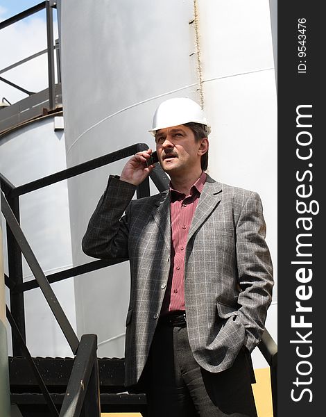 Engineer talking in phone, wearing hard-hat, on background with storage tank. Engineer talking in phone, wearing hard-hat, on background with storage tank