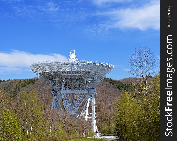 Observatory with large complicated metal dish and support structure surrounded by trees and forest, bright blue sky with a few clouds. Observatory with large complicated metal dish and support structure surrounded by trees and forest, bright blue sky with a few clouds.