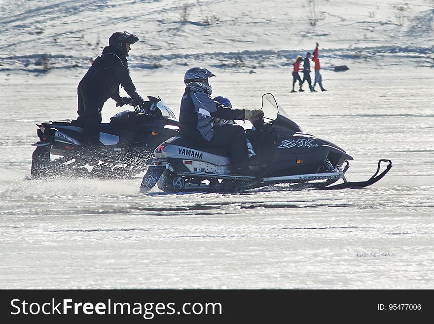 A pair of snow mobiles with people riding them in the snow. A pair of snow mobiles with people riding them in the snow.