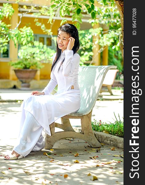 Young smiling Asian girl wearing white dress sitting in the park posing, blurred background. Young smiling Asian girl wearing white dress sitting in the park posing, blurred background.