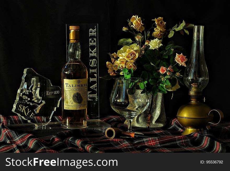 Table covered in tartan cloth with bottle of Talisker whisky vase of cream and pink roses, oil lamp, glass and map of Isle of Skye in glass, dark background. Table covered in tartan cloth with bottle of Talisker whisky vase of cream and pink roses, oil lamp, glass and map of Isle of Skye in glass, dark background.