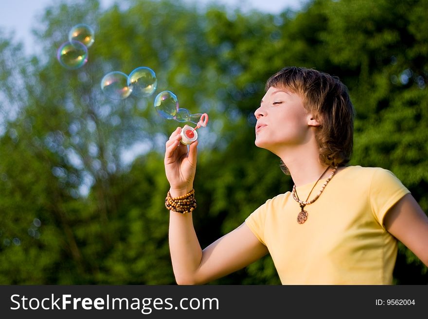 A young woman blowing up soap-bubbles on the green trees background. A young woman blowing up soap-bubbles on the green trees background