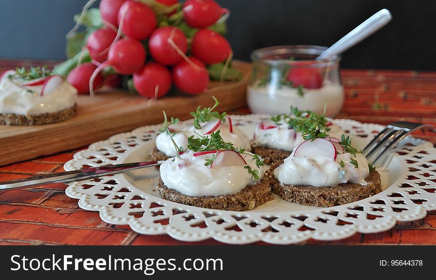 Four slices of baked bread with white sauce and garnishment on a white plate placed on a table, with a single bread and sauce, radishes, and a bowl of sauce in the background. Four slices of baked bread with white sauce and garnishment on a white plate placed on a table, with a single bread and sauce, radishes, and a bowl of sauce in the background.