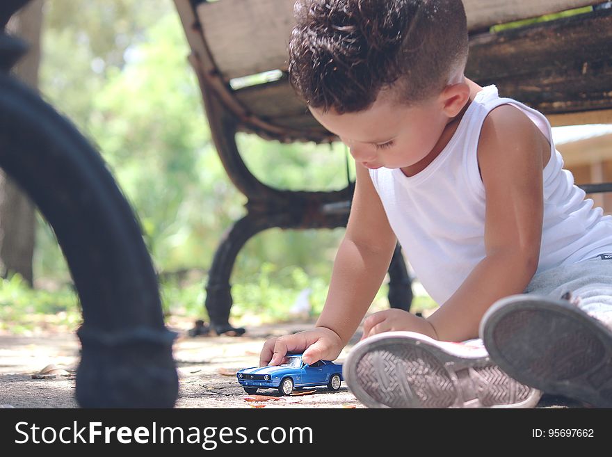 Boy in White Tank Top Playing Blue Coupe Die Cast Near Brown Wooden Bench Chair during Daytime