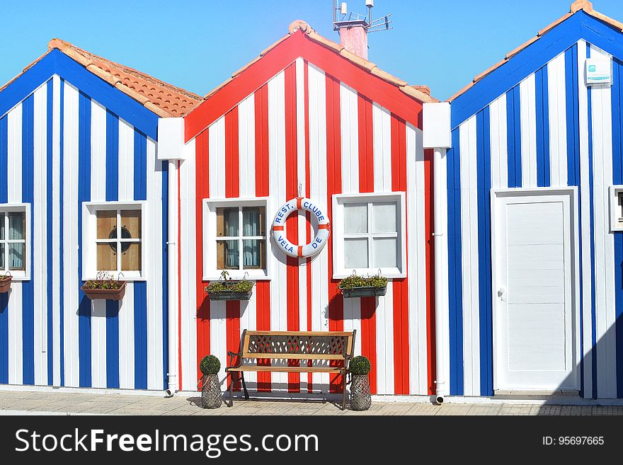 Three red and blue striped beach huts with tiled roofs decorated with a lifebelt and with a bench seat in front, blue sky and sunshine. Three red and blue striped beach huts with tiled roofs decorated with a lifebelt and with a bench seat in front, blue sky and sunshine.