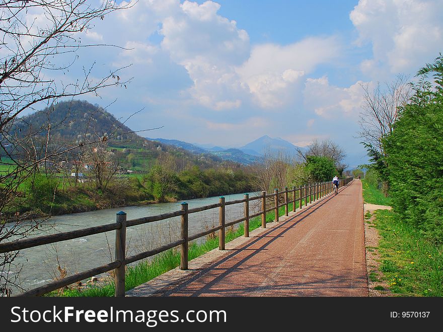 Italy. A valley of the mountain river. The Alpes are visible. Solar spring day. Along small river â€“ a new bicycle path. Far - a leaving bicyclist. Italy. A valley of the mountain river. The Alpes are visible. Solar spring day. Along small river â€“ a new bicycle path. Far - a leaving bicyclist.