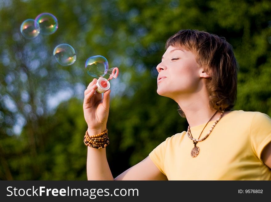 A young woman blowing up soap-bubbles on the green trees background. A young woman blowing up soap-bubbles on the green trees background