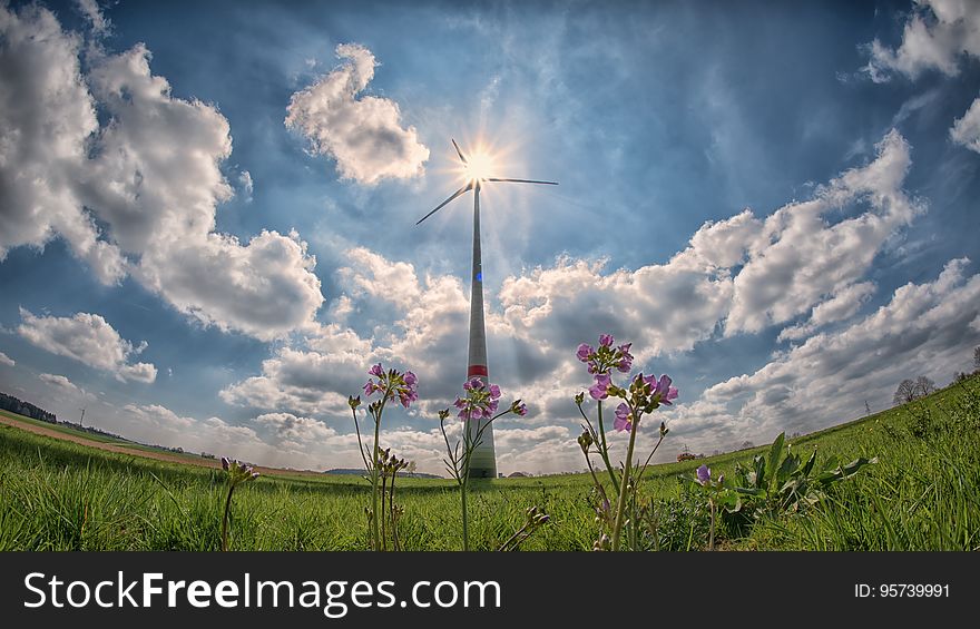 Fish eye view of wind turbine in countryside field with sun shining through blades and cloudscape background.