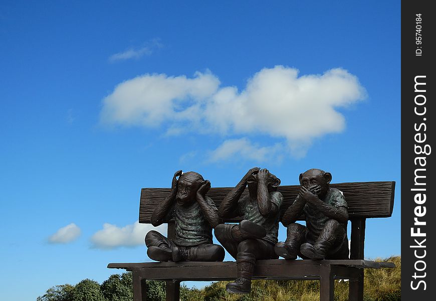 Three little brown monkeys, sculpted from wood, on a wooden bench seat in a park, background blue sky and cloud. The subject could be 'See no evil, hear no evil and speak no evil'. Three little brown monkeys, sculpted from wood, on a wooden bench seat in a park, background blue sky and cloud. The subject could be 'See no evil, hear no evil and speak no evil'.