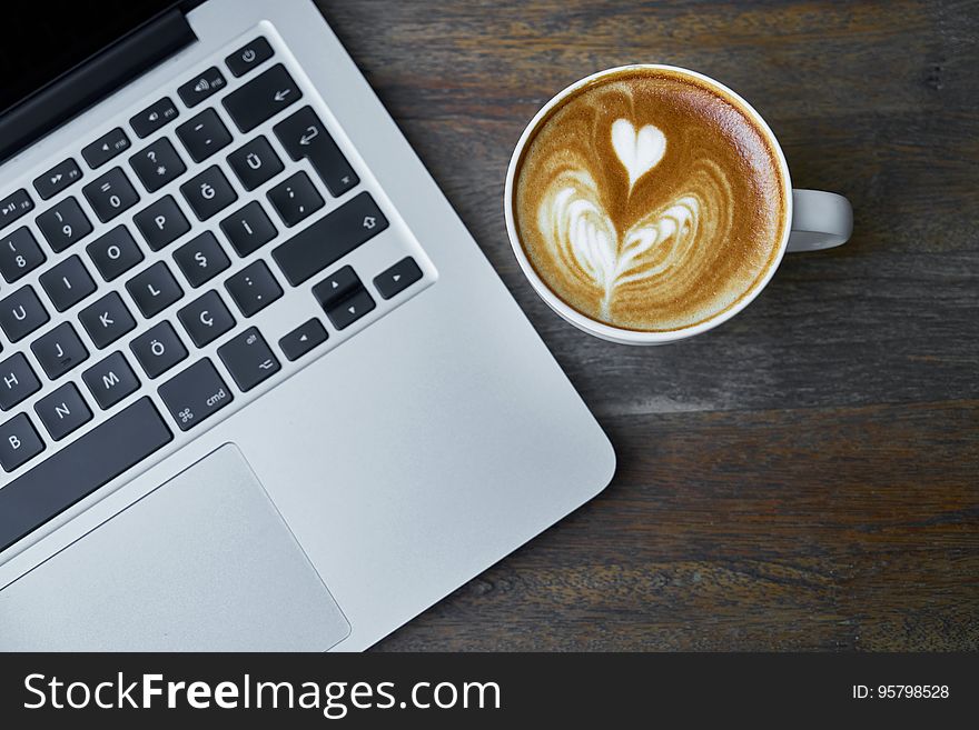 Coffee mug with heart shape in foam next to open laptop computer. Coffee mug with heart shape in foam next to open laptop computer.