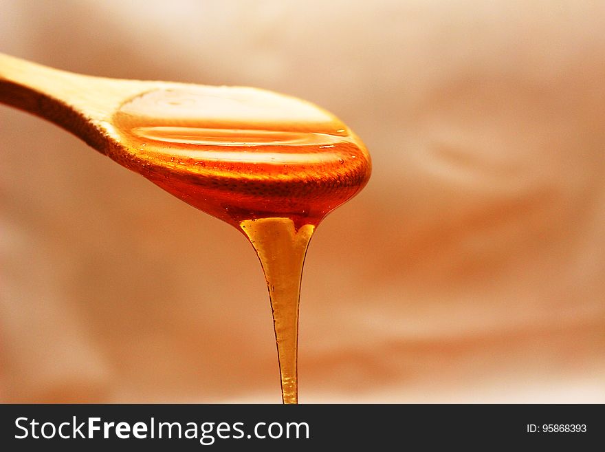 Spoonful of honey with thick sweet liquid flowing over the brim, pale gold background.
