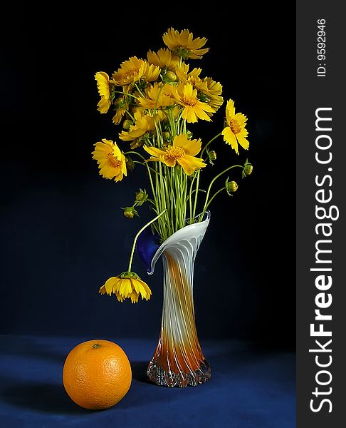 Bouquet of yellow flowers in a vase and an orange against a dark background