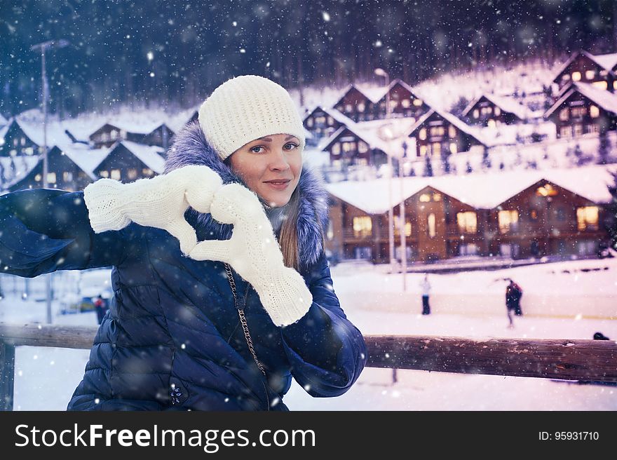 Portrait of woman in winter coat and cap making heart shape with mittens on hands outdoors in snowy town. Portrait of woman in winter coat and cap making heart shape with mittens on hands outdoors in snowy town.