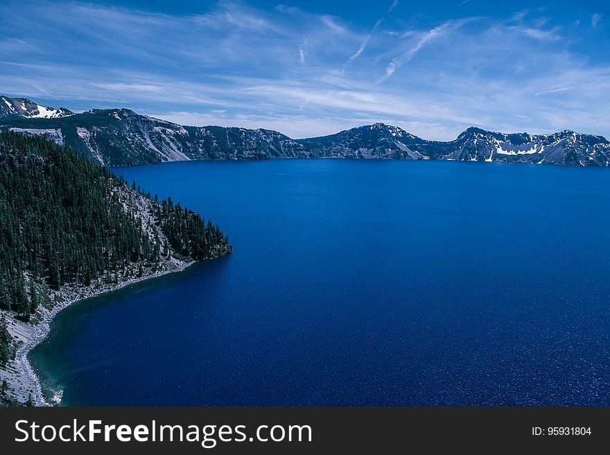 Aerial view of blue crater lake with pine tree forest in snow against blue skies on sunny day.